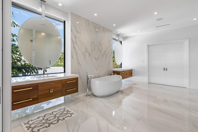 bathroom with stone wall, two vanities, a sink, marble finish floor, and a soaking tub