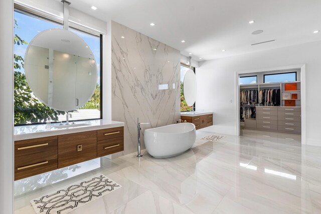 bathroom featuring a soaking tub, a sink, marble finish floor, stone wall, and two vanities