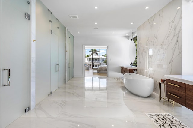 ensuite bathroom with connected bathroom, marble finish floor, a freestanding bath, vanity, and stone wall