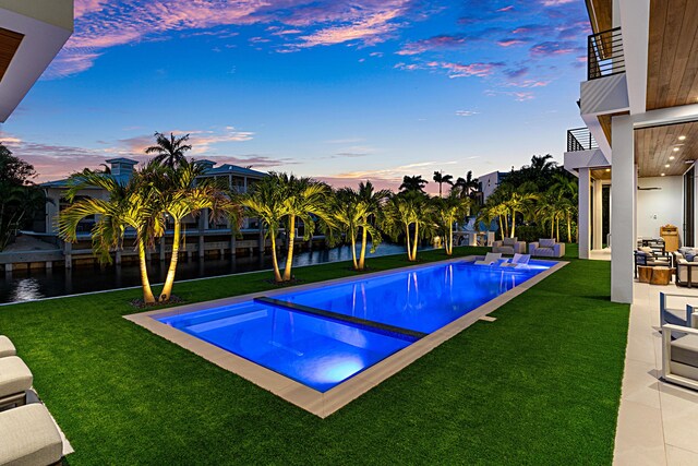 pool at dusk with a water view, a patio area, an outdoor pool, and a yard