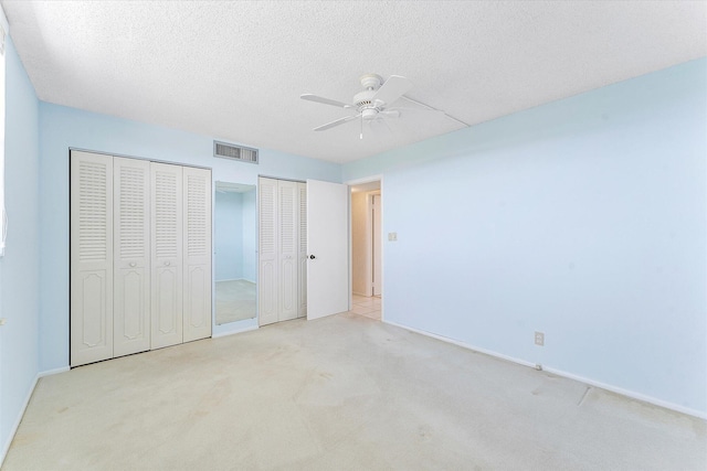 unfurnished bedroom with a textured ceiling, light colored carpet, a ceiling fan, visible vents, and two closets