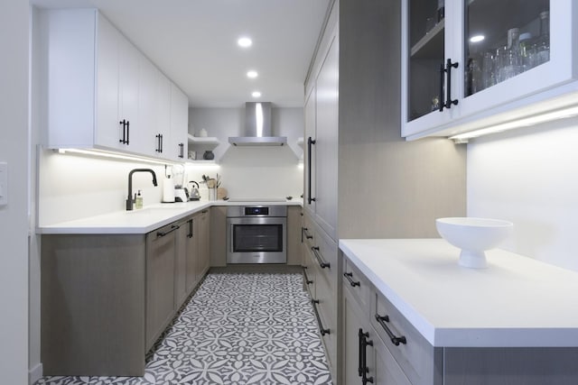 kitchen with light countertops, wall chimney range hood, stainless steel oven, open shelves, and a sink