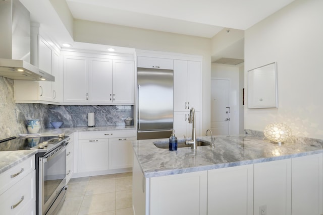 kitchen featuring a sink, light stone counters, appliances with stainless steel finishes, a peninsula, and wall chimney range hood