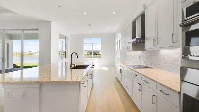 kitchen with a center island with sink, black electric cooktop, light wood-type flooring, wall chimney range hood, and a sink