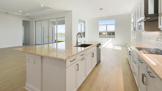 kitchen with light wood finished floors, wall chimney exhaust hood, a kitchen island with sink, stainless steel appliances, and a sink