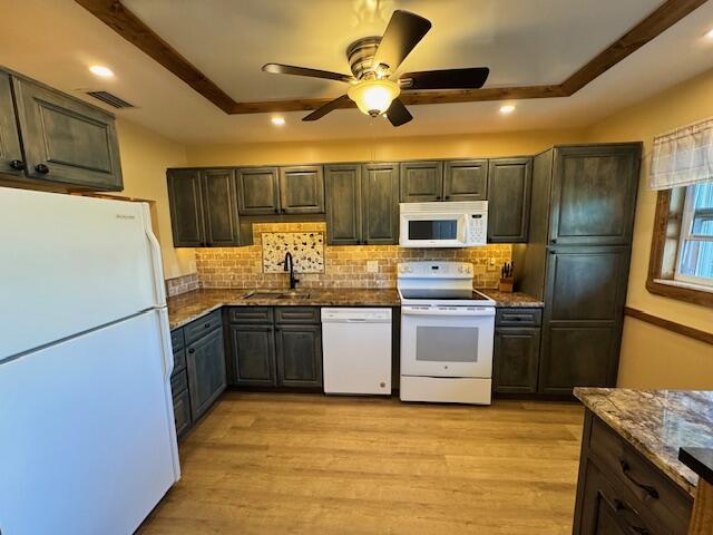 kitchen with dark stone countertops, light wood-style floors, white appliances, a raised ceiling, and a sink