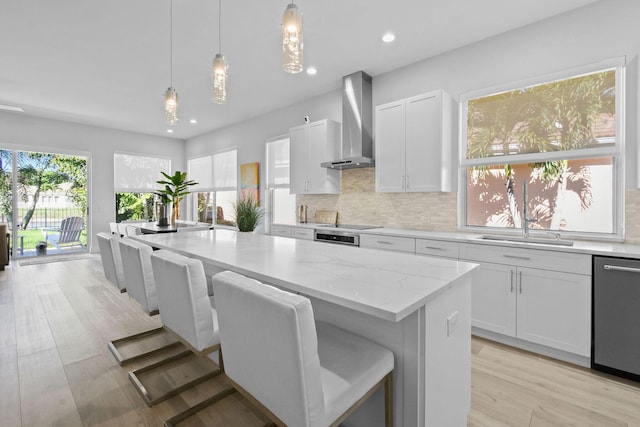 kitchen with a sink, backsplash, stainless steel dishwasher, white cabinets, and wall chimney range hood