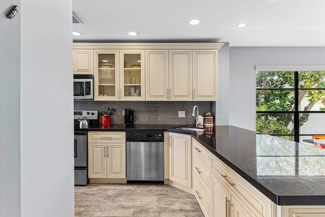 kitchen with a peninsula, a sink, decorative backsplash, stainless steel appliances, and cream cabinetry