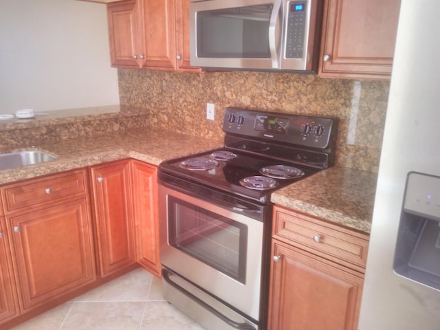 kitchen with brown cabinets, appliances with stainless steel finishes, light tile patterned floors, decorative backsplash, and light stone countertops