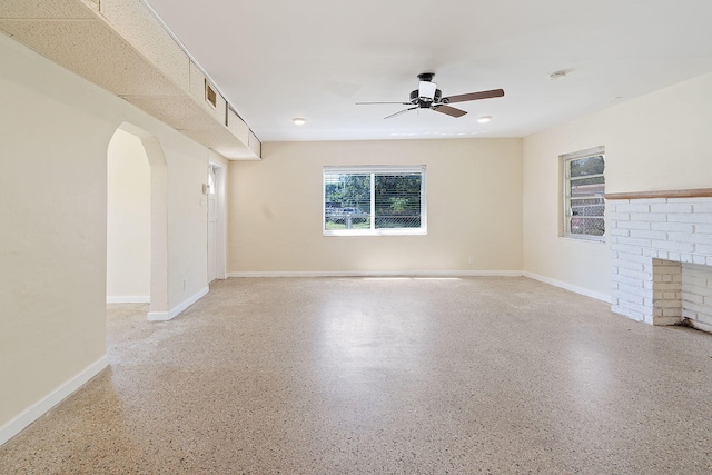 unfurnished living room with arched walkways, baseboards, ceiling fan, a brick fireplace, and light speckled floor