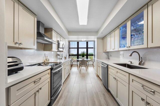 kitchen with a sink, appliances with stainless steel finishes, wall chimney exhaust hood, light wood finished floors, and tasteful backsplash