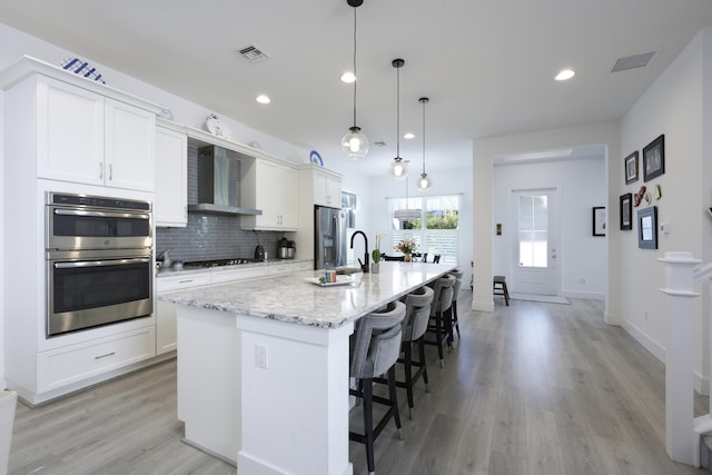 kitchen with tasteful backsplash, visible vents, appliances with stainless steel finishes, wall chimney range hood, and an island with sink