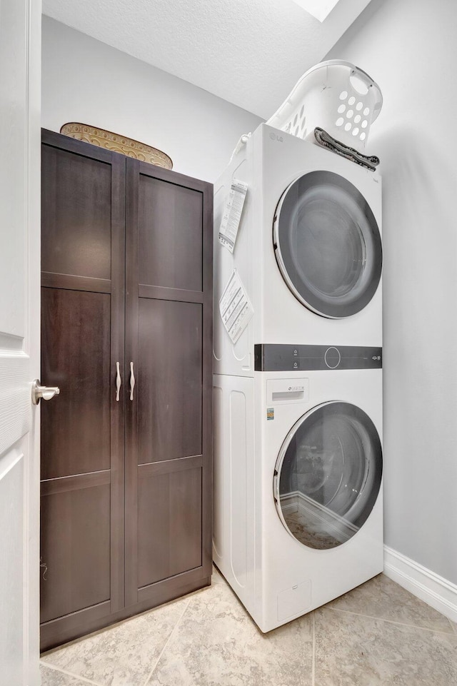 laundry room with a textured ceiling, cabinet space, light tile patterned floors, stacked washer / drying machine, and baseboards