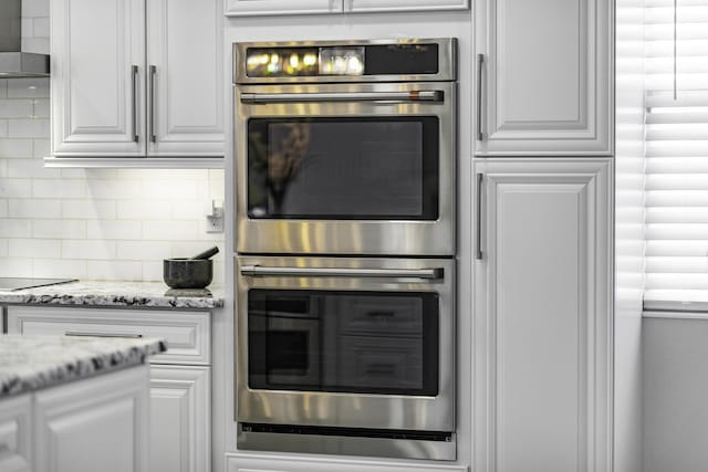 kitchen with decorative backsplash, double oven, wall chimney exhaust hood, and white cabinets