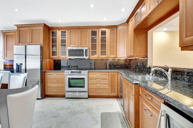 kitchen with stainless steel appliances, backsplash, glass insert cabinets, a sink, and dark stone countertops