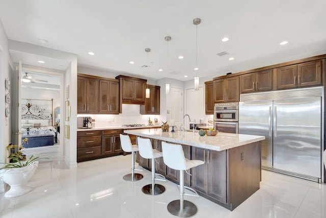 kitchen featuring light tile patterned floors, an island with sink, appliances with stainless steel finishes, decorative light fixtures, and backsplash