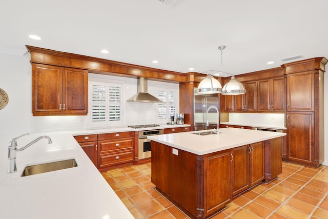 kitchen featuring stainless steel appliances, light countertops, a sink, wall chimney range hood, and an island with sink