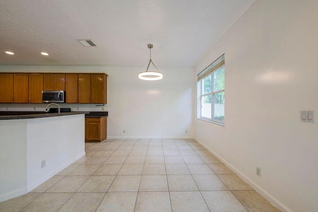 kitchen with light tile patterned floors, stainless steel microwave, visible vents, brown cabinetry, and dark countertops