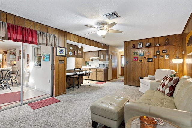 living room featuring wooden walls, visible vents, ceiling fan, a textured ceiling, and light colored carpet