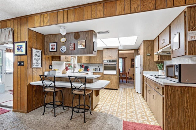 kitchen featuring white appliances, wooden walls, a peninsula, light countertops, and under cabinet range hood