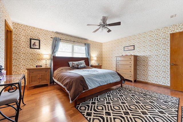 bedroom featuring baseboards, wallpapered walls, ceiling fan, a textured ceiling, and light wood-type flooring