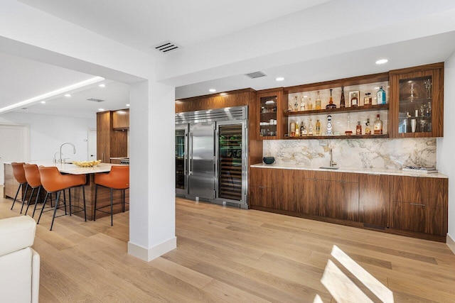 bar with visible vents, indoor wet bar, light wood-type flooring, backsplash, and stainless steel built in refrigerator