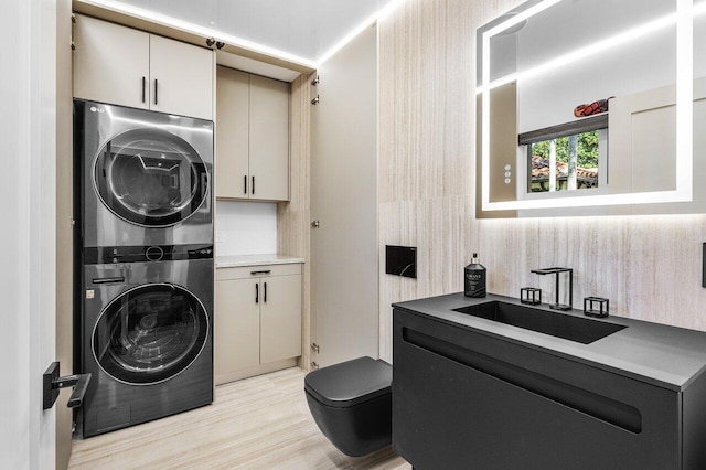 clothes washing area with light wood finished floors, cabinet space, stacked washing maching and dryer, and a sink