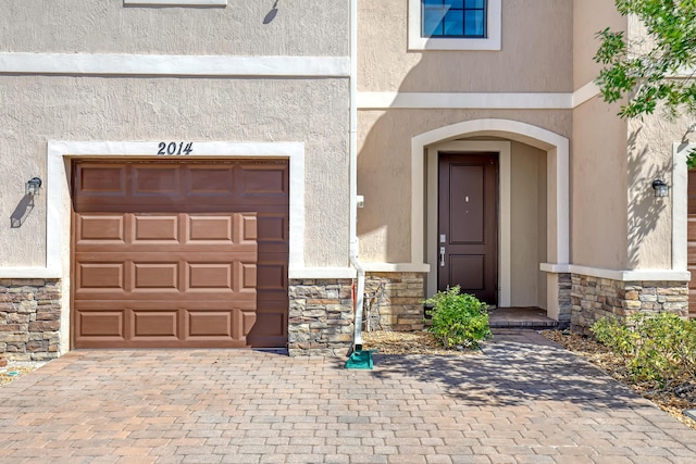 entrance to property with a garage, stone siding, decorative driveway, and stucco siding