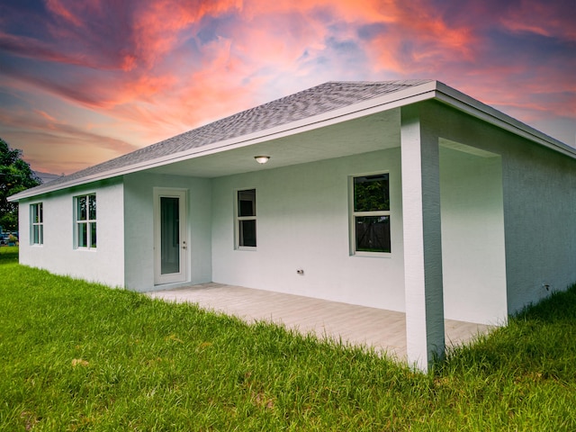 back of house at dusk with a shingled roof, a lawn, a patio, and stucco siding