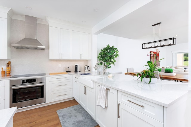 kitchen with oven, light wood-type flooring, a peninsula, wall chimney range hood, and black electric cooktop