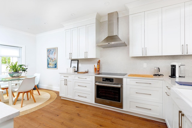 kitchen with ornamental molding, oven, light wood-style floors, wall chimney range hood, and black electric cooktop