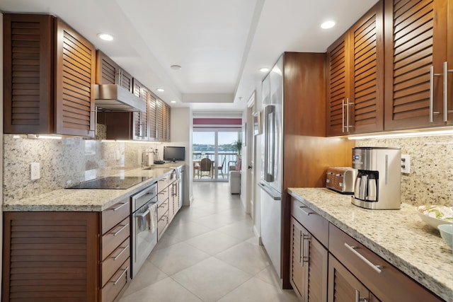 kitchen featuring light stone counters, oven, black electric cooktop, under cabinet range hood, and a tray ceiling