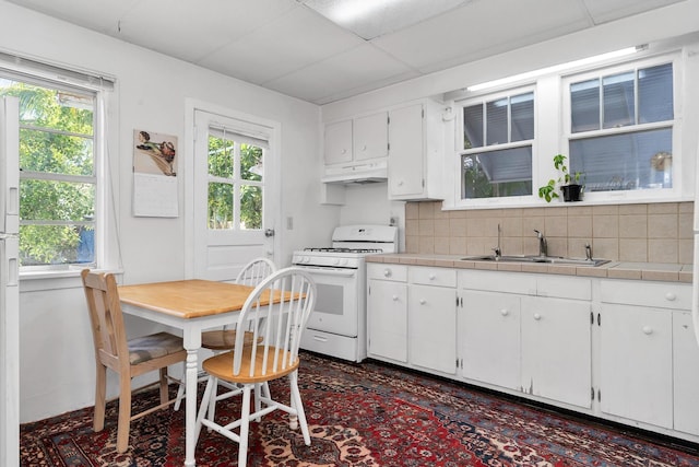 kitchen with white gas stove, under cabinet range hood, a sink, backsplash, and tile countertops