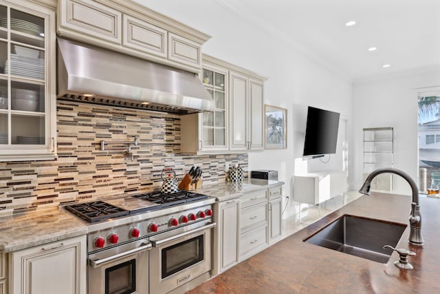 kitchen featuring wall chimney range hood, double oven range, a sink, cream cabinets, and backsplash