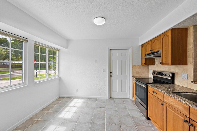 kitchen featuring brown cabinets, electric range, decorative backsplash, dark stone counters, and under cabinet range hood