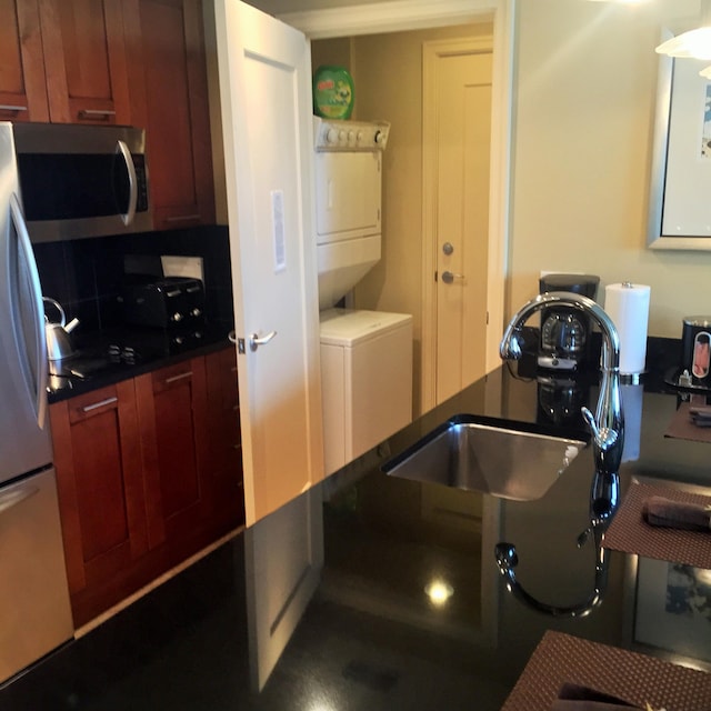 kitchen featuring a sink, stainless steel microwave, dark countertops, freestanding refrigerator, and stacked washer / dryer