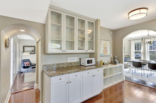 kitchen with white microwave, light stone countertops, arched walkways, glass insert cabinets, and white cabinetry