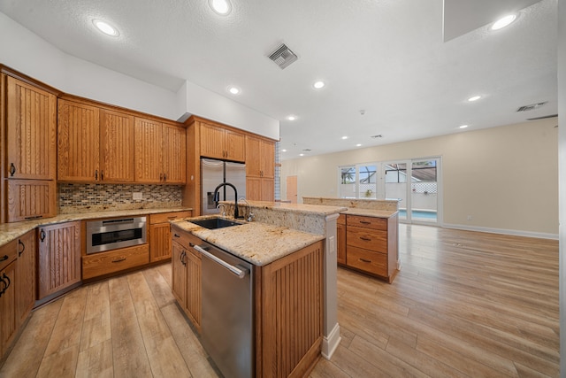 kitchen featuring light wood-style flooring, a kitchen island with sink, a sink, backsplash, and appliances with stainless steel finishes