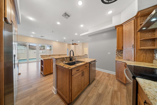 kitchen featuring light wood-style flooring, an island with sink, a sink, appliances with stainless steel finishes, and wall chimney range hood