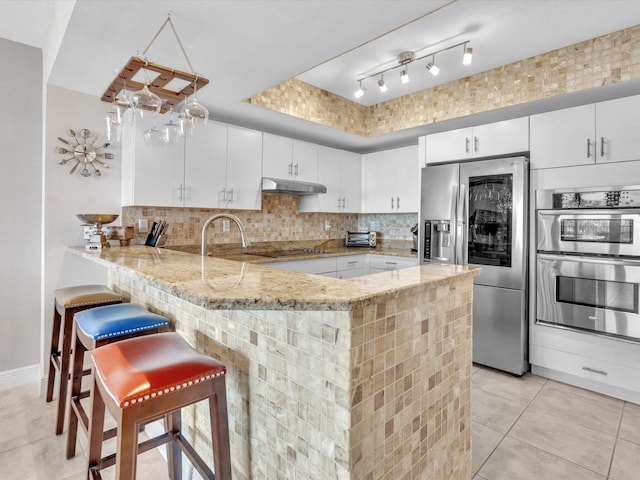 kitchen featuring light stone countertops, under cabinet range hood, decorative backsplash, appliances with stainless steel finishes, and a peninsula