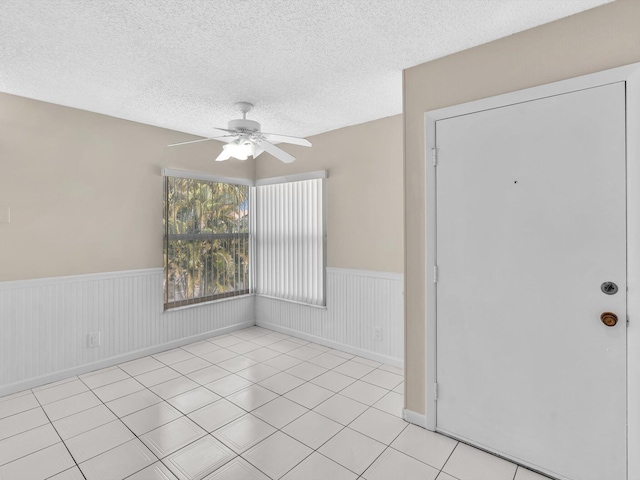 empty room with a textured ceiling, light tile patterned floors, a ceiling fan, and wainscoting