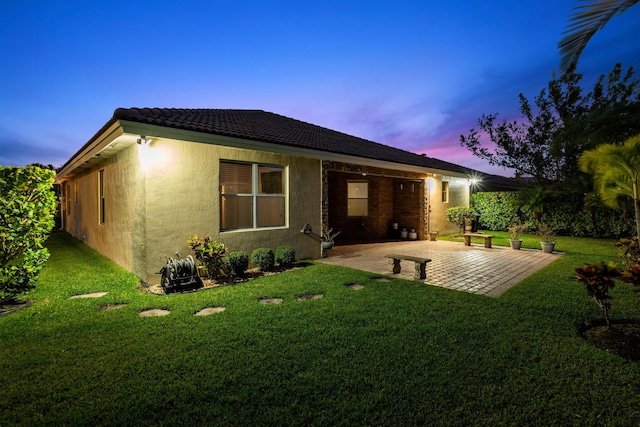 back of house at dusk with a yard, stucco siding, a tiled roof, and a patio