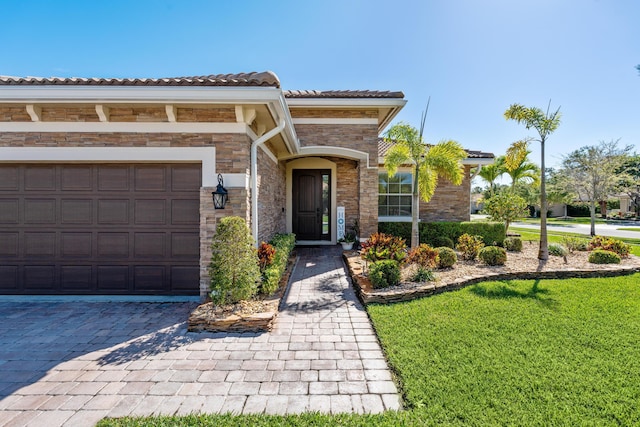 entrance to property featuring decorative driveway, stone siding, a yard, an attached garage, and a tiled roof