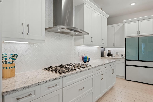 kitchen featuring decorative backsplash, wall chimney range hood, stainless steel gas stovetop, white cabinetry, and built in fridge