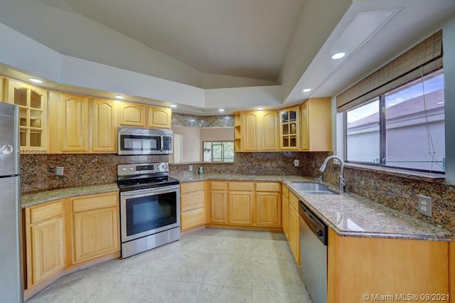 kitchen featuring light brown cabinetry, lofted ceiling, light stone counters, stainless steel appliances, and a sink