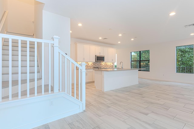 kitchen featuring a center island with sink, visible vents, decorative backsplash, stainless steel microwave, and white cabinetry