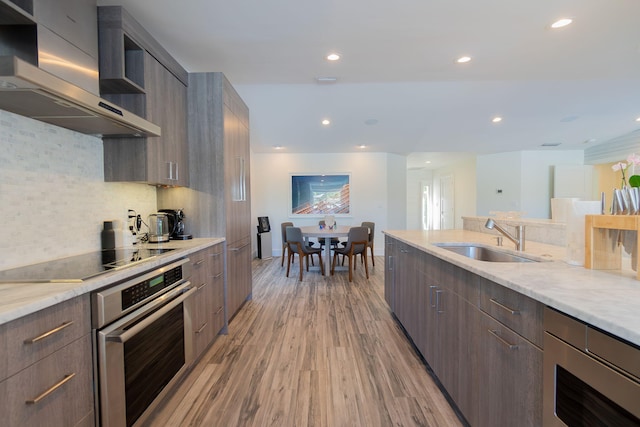 kitchen featuring modern cabinets, a sink, wall chimney range hood, stainless steel oven, and black electric cooktop