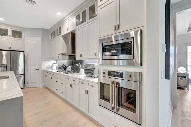 kitchen featuring tasteful backsplash, glass insert cabinets, light wood-style flooring, stainless steel appliances, and wall chimney exhaust hood