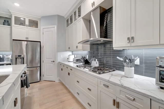 kitchen featuring white cabinets, light wood-style floors, appliances with stainless steel finishes, wall chimney range hood, and tasteful backsplash