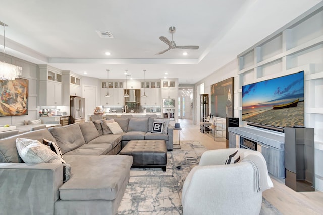 living room with light wood finished floors, visible vents, a tray ceiling, recessed lighting, and ceiling fan with notable chandelier